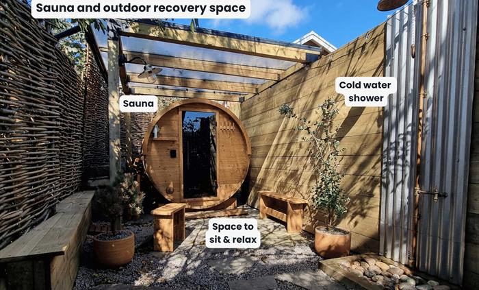 An outdoor, round wooden barrel sauna tucked away under the trees with an outdoor shower and benches for sitting on